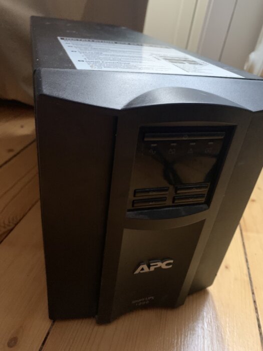 En APC UPS (uninterruptible power supply) enhet på ett trägolv. Dustig, svart, backup strömförsörjning.