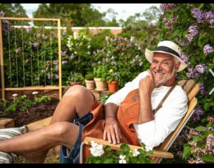 En leende man i hatt vilar på en stol i en blomstrande trädgård.