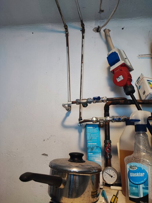 Rörledningar och mätinstrument på vägg, rostfri gryta och städprodukter på arbetsyta, inomhusmiljö.