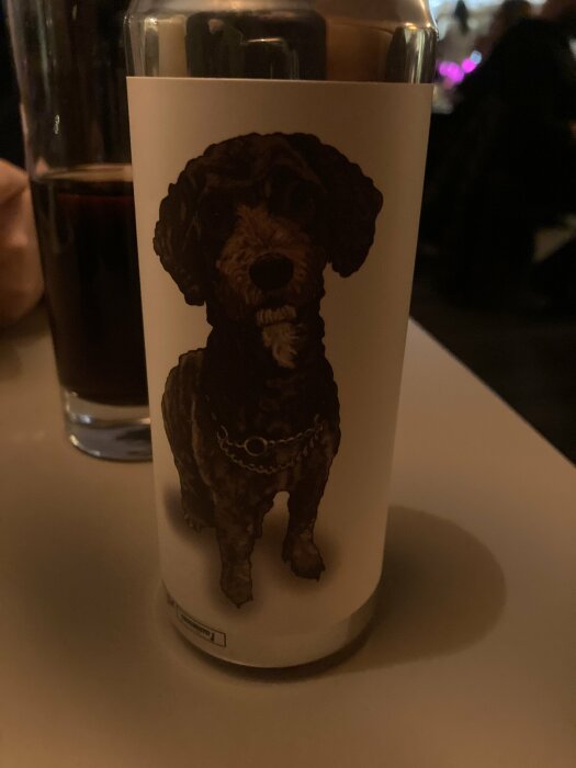 Ett ljus med hundmotiv framför en out-of-focus bakgrund med dunkelt ljus och ett mörkt glas dryck.