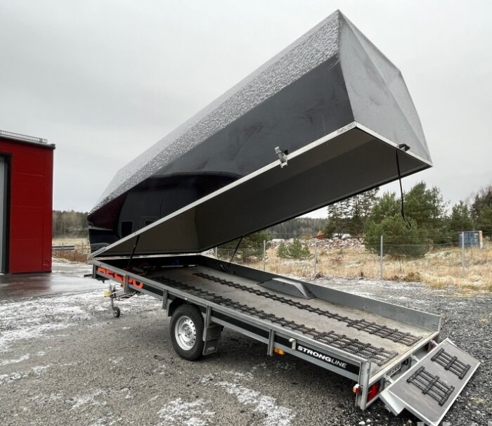 Släpvagn med tippfunktion och öppet flak, parkerad utomhus, snöfläckar på marken, molnig himmel.