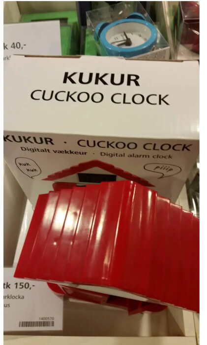 Förpackning för röd "KUKUR CUCKOO CLOCK", prisinformation, omgivande föremål suddigt i bakgrunden.