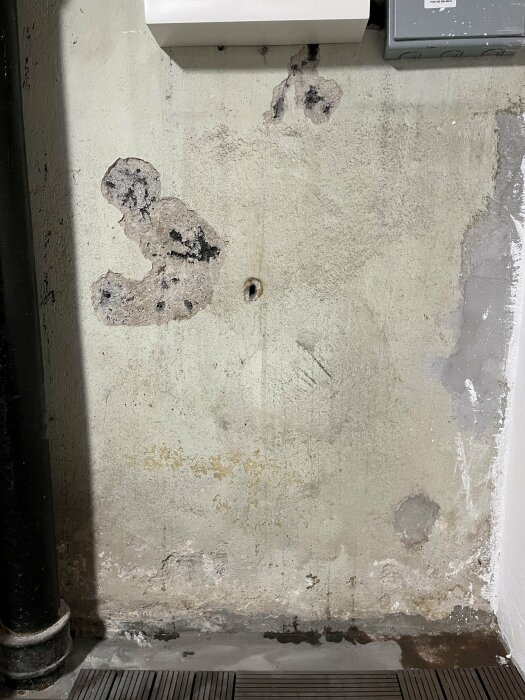 Vägg med flagnande färg, smuts, rör och en elektrisk enhet. Slitet och möjligen fuktproblem.