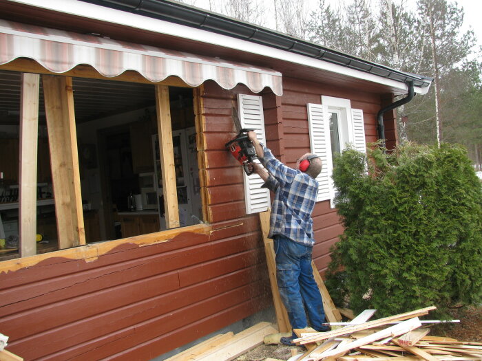 Person bygger om hus, använder borr, bär hörselskydd, lantlig miljö, trävägg, trädgårdsarbete.