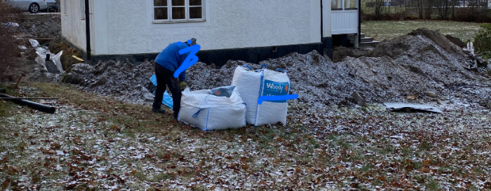 Person i blått arbetskläder vid säckar, gråmulet väder, byggnadsarbete, högar av grus, frostigt gräs.