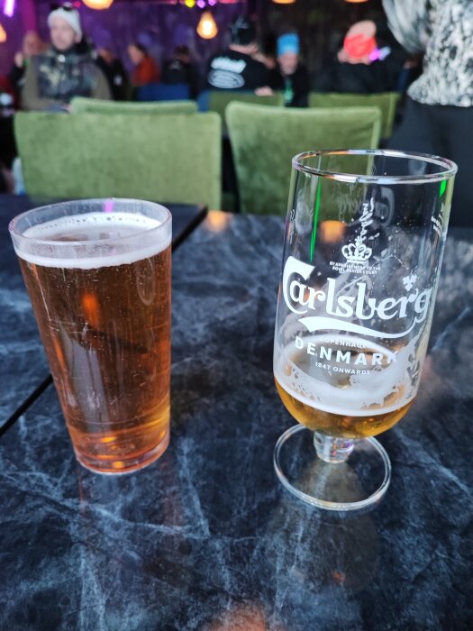 Två ölglas på bord, ett fullt, ett halvtomt. Bakgrund suddig, människor, inomhus miljö, festlig atmosfär.