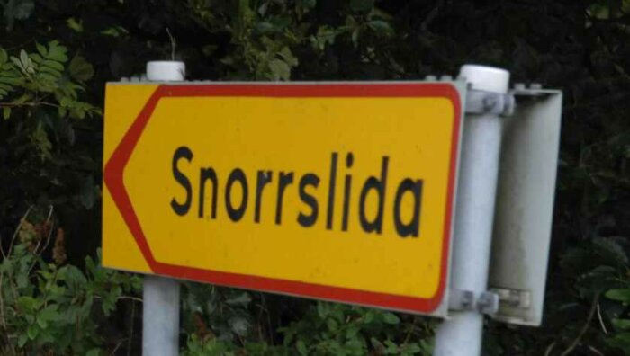 Gul och röd vägskylt med svart text "Snorrsliða", pil vänster mot grönbeklädd bakgrund.