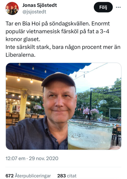 Man med glas öl utomhus på kvällen; belysning och vegetation i bakgrunden. (Tweet med text synlig.)