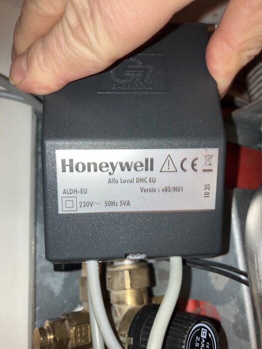 Ett handtag håller en elektrisk komponent med etiketter och varningsymboler. Märke: Honeywell. Rör och metall i bakgrunden.