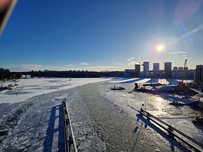Vinterlandskap, solig, isbelagd flod, båtar, bro, moderna byggnader, konstruktion, skuggor, blå himmel, kallt väder.