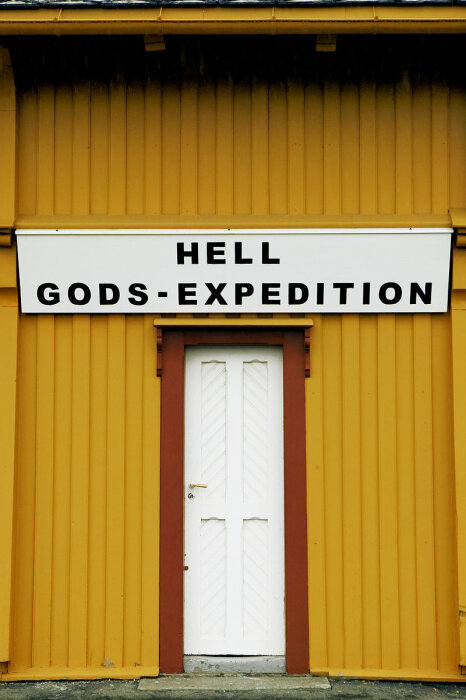 Gul vägg, vit dörr, skylt med text "HELL GODS-EXPEDITION", arkitektoniska detaljer, ingång, humoristiskt ordspel.