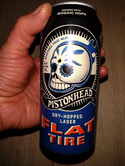 Hand håller en ölburk med dödskalle-design, märket "Pistonhead", "Flat Tire" Dry-Hopped Lager, 4,5% alkohol.