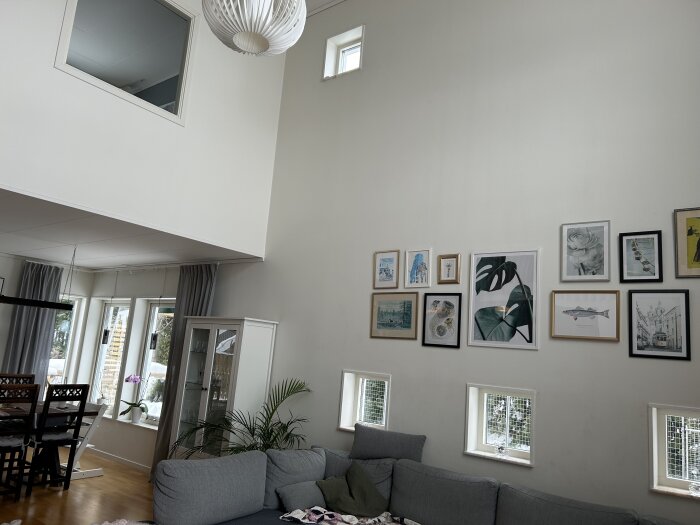 Ljus vardagsrum, högt i tak, grå soffa, konst på vägg, taklampa, fönster, gröna växter, speglingen av ett rum ovanpå.