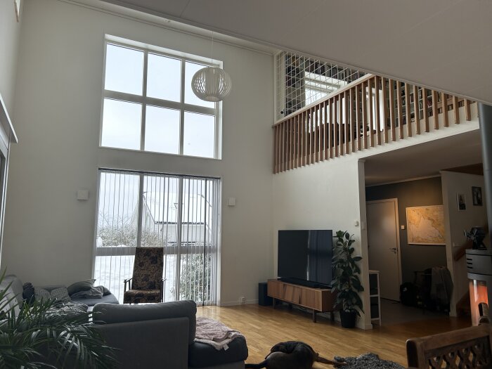 Vardagsrum med högt i tak, stort fönster, loft, TV, soffa, växt, hund, snö utanför.