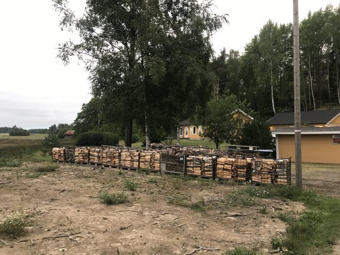 Staplar av ved i förgrunden, gula hus och träd i bakgrunden, grå himmel, ingen snö, sannolikt sent på dagen.