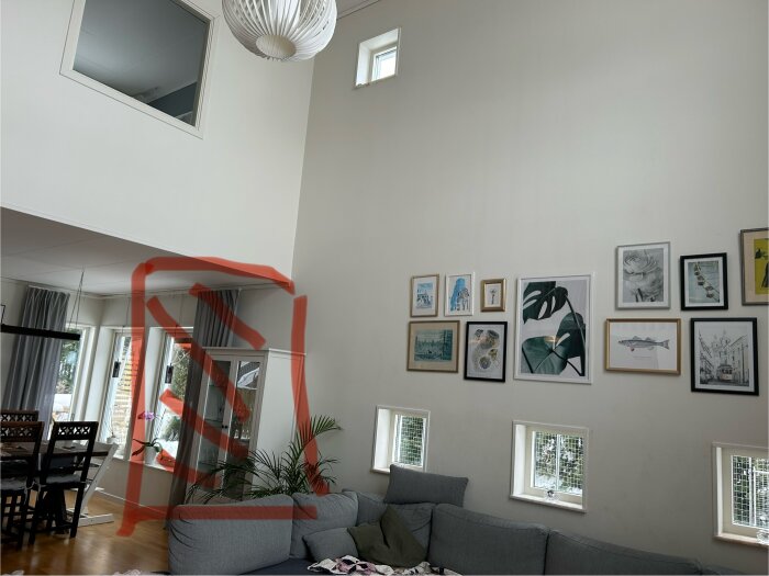 Modernt vardagsrum, högt i tak, vägg med konstinramningar, fönster, grå soffa, röd markering ritar upp något.