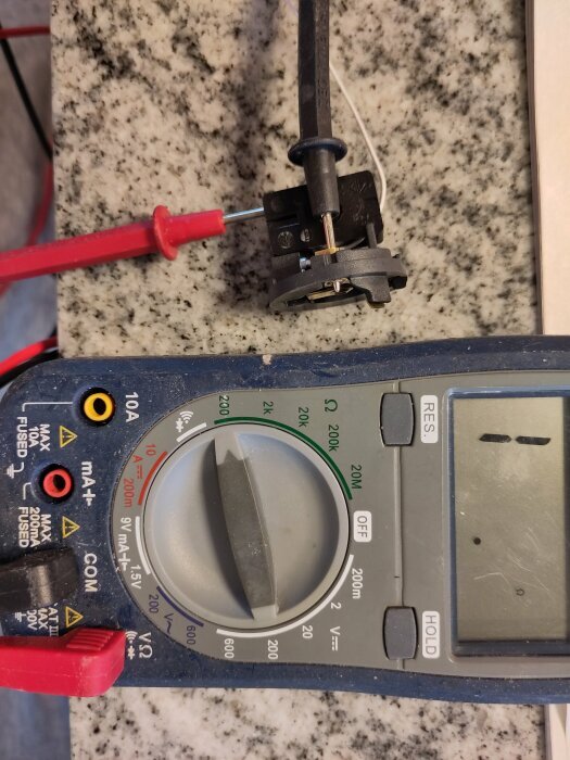 Multimeter mäter komponent, förmodligen motstånd, på arbetsyta, testkabel ansluten, teknisk, elektronikreparation.