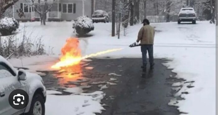 Person använder eldkastare för att smälta snö på uppfart, i snöigt bostadsområde.