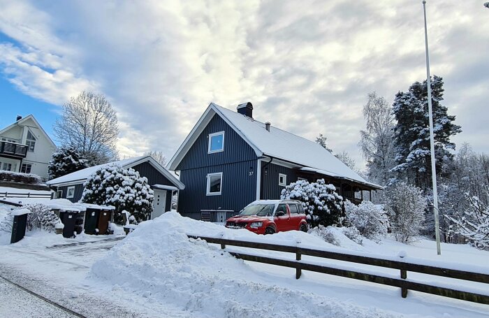 Ett blått hus, snötäckt landskap, röd bil, vinterdag, molnigt, kala träd, bostadsområde.