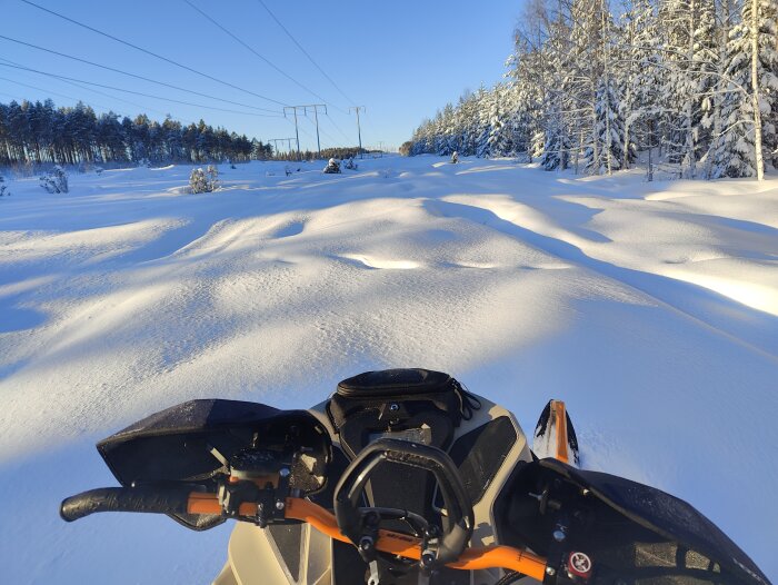 Snöfält, skoterstyre, skog, soligt, vinterdag, strömförande ledningar i bakgrunden.