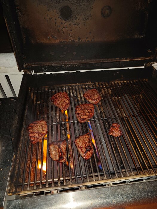 Stekta köttbitar på en grill med synliga lågor och grillränder. Använd, smutsig grill inomhus eller utomhus.