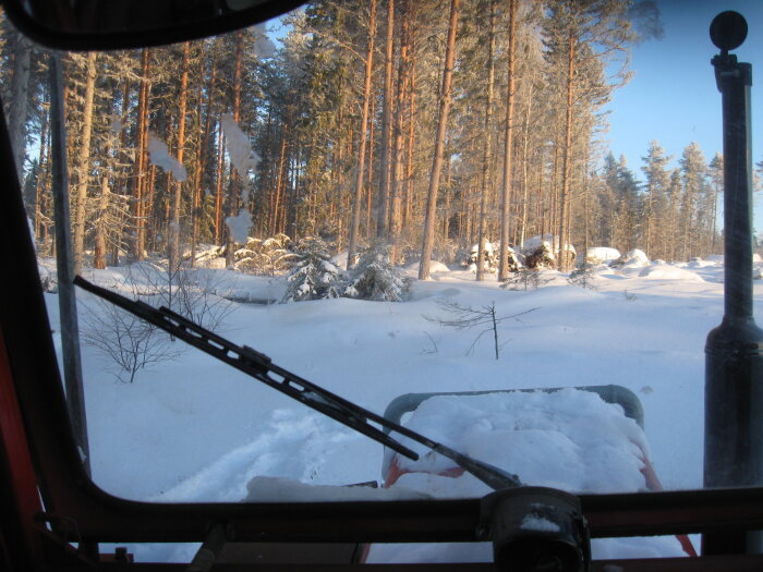 Vy genom snöig terräng från insidan av en fordonshytt. Soligt väder, tallskog, snödrivor och ett vindrutetorkare.