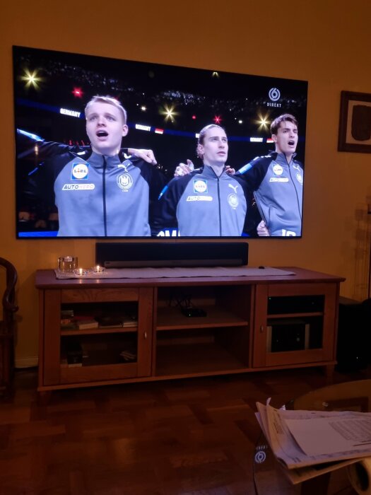 Tre unga män sjunger nationalsången på sportevenemang på TV i ett vardagsrum.