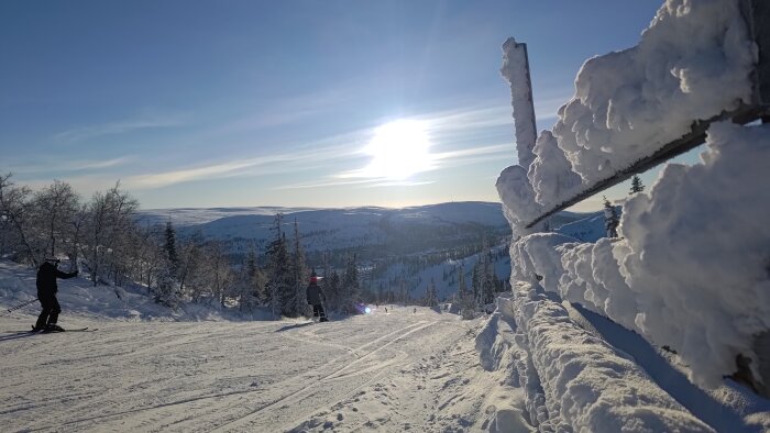 Skidbacke med snöklädda träd, skidåkare, solig himmel, tjock snö på trästaket.