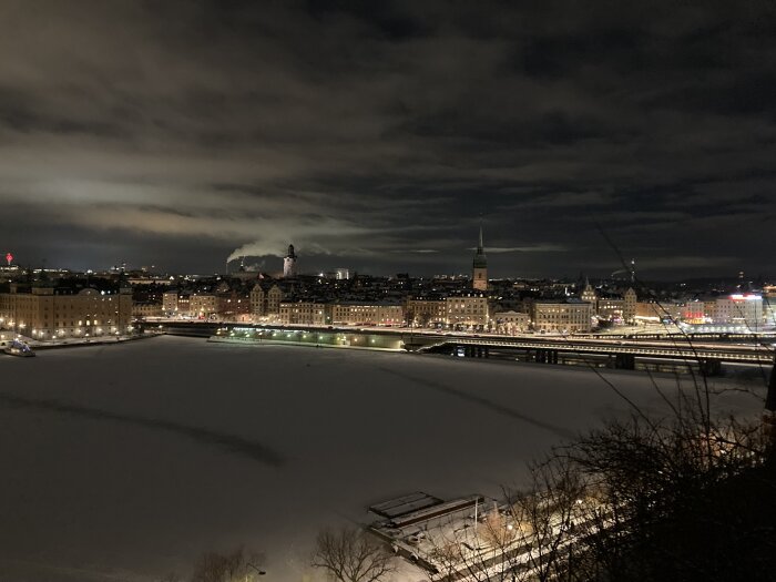 Stadssilhuett vid natt, belysta byggnader, isbelagt vatten, molnigt, vinter, bro, reflektioner, urban atmosfär.