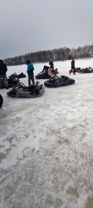 Personer i vinterkläder, iskörning med go-karts på en frusen sjö, grå himmel, skogsbakgrund.