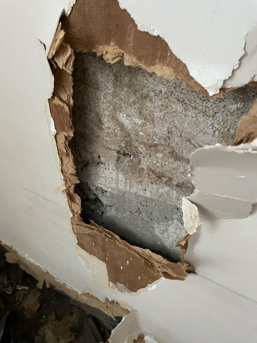 En skadad vägg med avslöjat betong och framtornande lager av gipsskivor eller väggmaterial. Reparation behövs.