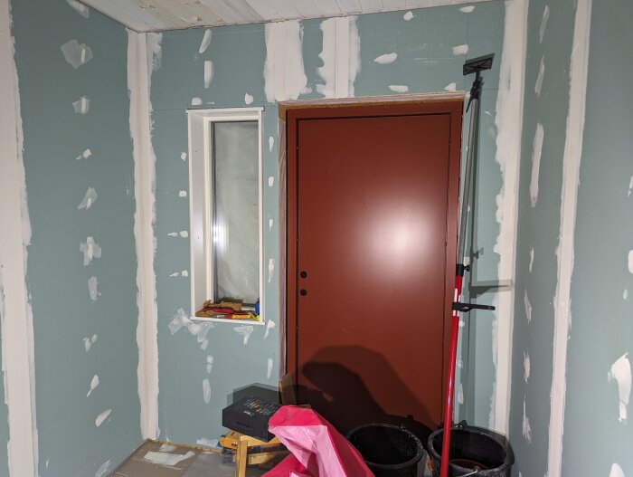 Ett rum under renovering med spacklade väggar, en brun dörr och fönster, städredskap synliga.