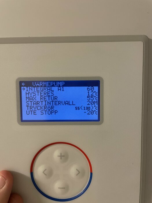 Digital display för värmepump med temperaturinställningar och knappar för navigering, omgiven av vit vägg.