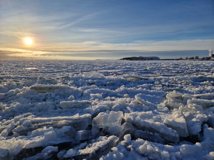 Solnedgång över isbelagt landskap, snötäckta isblock, öppet vatten i bakgrunden, klart väder, vinterstämning.