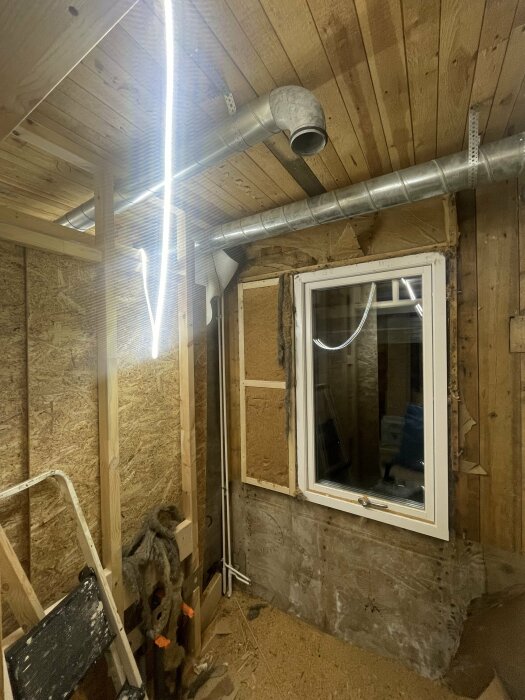 Inomhuskonstruktion under arbete, isoleringsmaterial, ventileringsrör, oskyddade väggar, träreglar, fönster, arbetsbelysning.