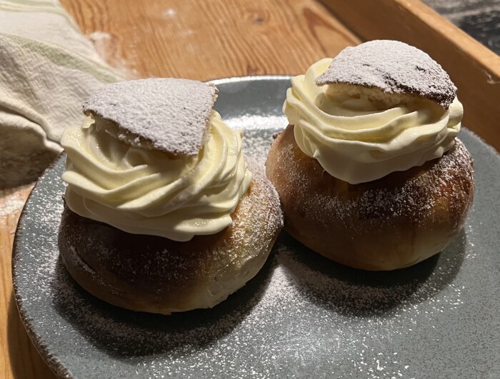 Två semlor med grädde och florsocker på ett grått fat.