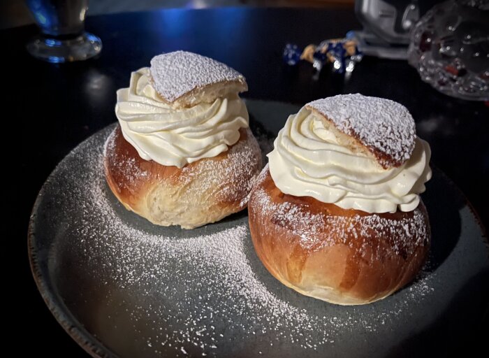 Två semlor med grädde och florsocker på mörk tallrik.