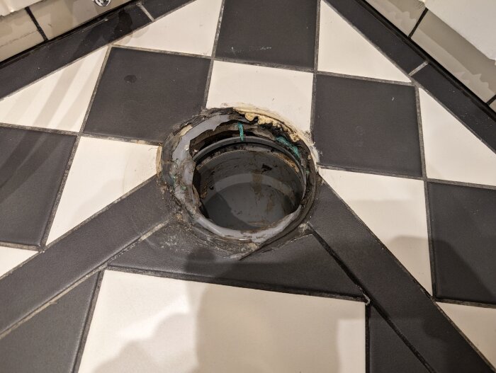 Öppet avloppsrör i svartvit kaklad golv, behöver reparation eller ny toalettinstallation.