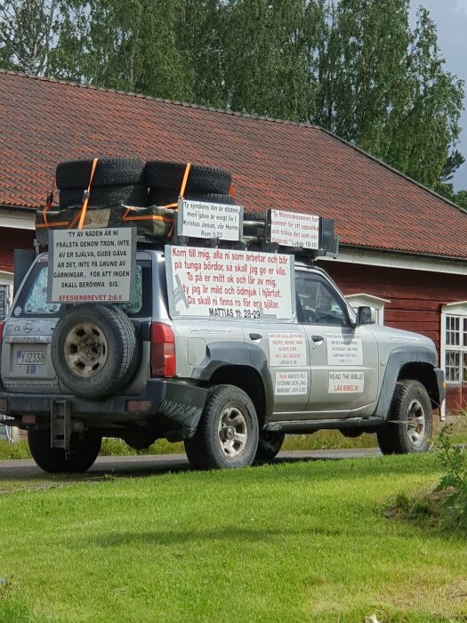 Terrängbil med reservdäck och religiösa budskap på skyltar parkerad framför hus.