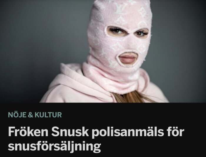 Person med maskering inomhus, uttrycksfull blick, rosa tröja, grå bakgrund, misstänkt för snusförsäljning.