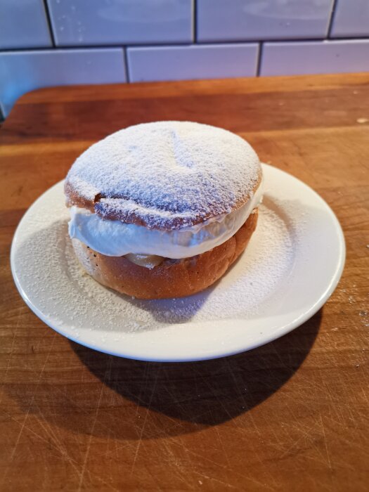 Semla på vit tallrik, florsocker ovanpå, gräddfyllning, träbord, kakel i bakgrunden.