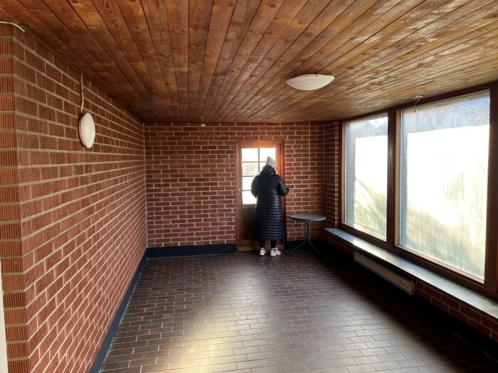 Tomt rum med tegelväggar, träbjälklag i taket, stora fönster, och en person står vid fönstret.