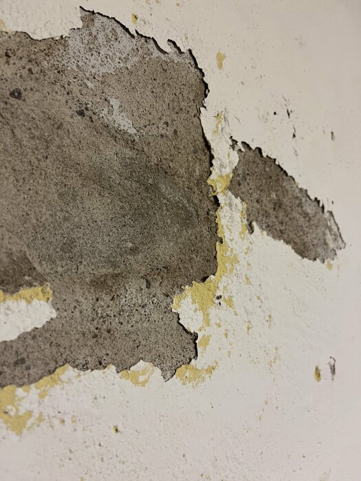 Skadad vägg med flagnande vit målfärg som avslöjar underliggande lager av gips och betong.