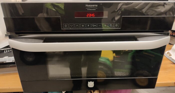 Modernt svart mikrovågsugn med digitala kontroller och klocka som visar 23:45. Reflektioner och föremål synliga inuti.
