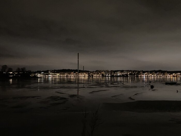 Nattvy, isbelagd sjö, stadssilhuett, reflekterande ljus, skymning, en skorsten, mörka moln.