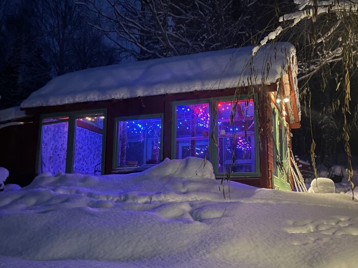 Snötäckt hus med färgglada ljus genom fönster; vinterkväll; lugn och mysig stämning.