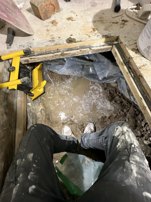 Person i smutsiga arbetskläder sitter vid ett öppet golv med vatten och verktyg synliga.