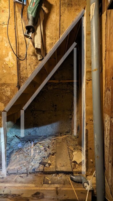 Rå byggnadsinredning, synliga träbjälkar, rör, skräp på golvet, ofärdiga trappsteg, borr på väggen, konstruktion eller renovering.