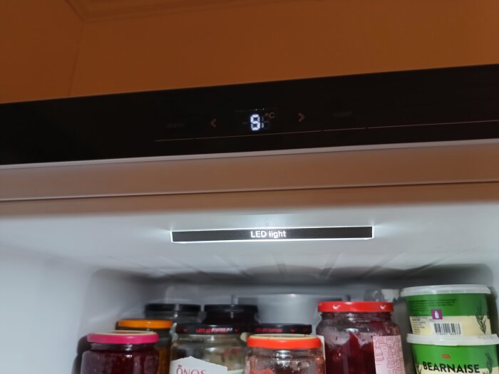 Digitalt kylskåpstermostat visar 9 grader Celsius, hylla med syltburkar och såsbehållare, LED-belysning.