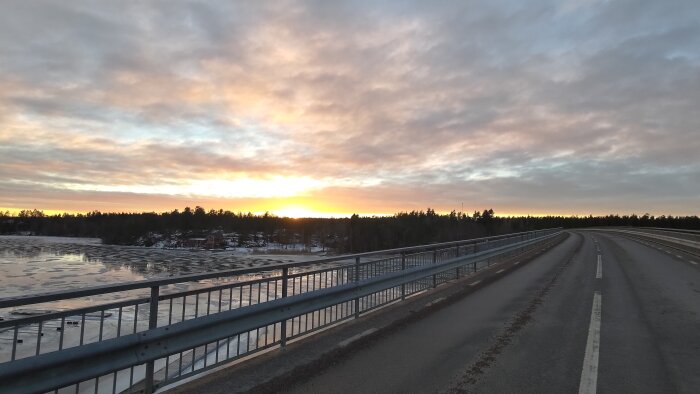 Väg över bro vid solnedgång, isbelagd sjö, träd, moln, lugn, kvällsljus, delvis isfritt vatten.